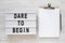 `Dare to begin` words on a lightbox, clipboard with blank sheet of paper on a white wooden surface, top view. Overhead, from abo