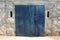 Dark blue dilapidated metal garage doors with rusted yellow frame mounted on stone wall with two openings for ventilation