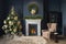 Dark blue living room with a fireplace and Christmas tree with gifts. A cozy armchair with a coffee table next to the fireplace. A