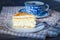 On a dark countertop, on a light checkered linen napkin, a white and blue patterned tea pair with tea and saucer with a piece of