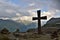 Dark cross on the mountain peak top with a number of small stones pyramids around - mountains and clouds are in the background