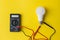 Dark Digital multimeter with probes and LED bulb on yellow background. Power saving concept. A multimeter is an electronic