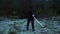 On a dark evening, a man in black ninja clothes stands with his back to the camera and holds a katana in his hand. The