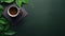 A dark green desk in an office with a closed paper notebook, green leaves of ornamental grass and a cup of coffee.