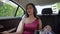 Dark-haired Asian young lady in pink dress is sitting in the backseat of the car and smiling, holding some carrier bags