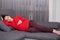 Dark haired attractive future mother lies on comfortable grey sofa, wearing warm bright red sweater and maroon leggings, holds her