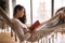 Dark-haired girl dressed in pants, sweater and warm slippers reads a book lying in a hammock in a cozy room with