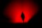 A dark, horror concept. Of a blurred hooded figure silhouetted against lights at night. With a black, red moody edit