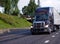 Dark modern big rig semi truck with grille guard and dry van semi trailer move with commercial cargo on straight wide highway road