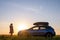 Dark silhouette of lonely woman relaxing near her car on grassy meadow enjoying view of colorful sunrise. Young female driver