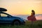 Dark silhouette of lonely woman relaxing near her car on grassy meadow enjoying view of colorful sunrise. Young female driver