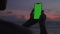 Dark silhouette man hold vertical mobile phone and watching to screen against background of colorful sunset. Use green