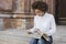 A dark-skinned girl sits on the steps and holds a tablet and looks into it . On his feet are books