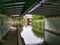 The darkened towpath of the Leeds Liverpool Canal as it passes under a rivetted metal bridge and pipework at the A5057