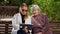 A daughter with a tablet and an elderly grandmother are sitting on a park bench.