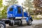 Day cab big rig blue semi truck tractor standing on the industrial parking lot with autumn trees in sunshine