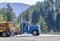 Day cab blue big rig semi truck transporting large wood logs on the semi trailer driving on the road with green forest