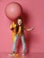 Dazed young lady, teen girl in jeans and yellow t-shirt with glass of yellow juice stands at big pink balloon with yellow tape