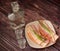 A decanter and two glasses of vodka on a wooden table, next to a plate with two sandwiches with bacon and green onions