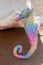 a decorative rainbow seahorse stands on the beach on a sunny day, near a large seashell