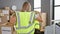 Dedicated young blonde woman volunteer standing with her back to us, wearing reflective vest in bustling charity center