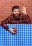Delicious concept. Man in checkered shirt near bottle, brick wall background. Barman with beard on cheerful face holds
