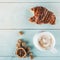 Delicious croissant with a bite on one side and hot cappuccino on blue wooden background, top view