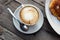 Delicious hot Italian cappuccino stands in a cup with a saucer on a wooden table. In the background a fresh croissant on a plate.