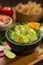 A delicious vertical shot Bowl of Guacamole next to fresh ingredients on a table with tortilla chips and salsa