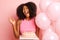 Delighted dark skinned ethnic woman, holds a cake with balloons, smiles expressively and laugh, isolated next to pink