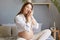 Delighted happy satisfied gentle pregnant woman with bare belly wearing white shirt sitting in bed in light bedroom embracing