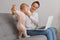 Delighted smiling dark haired woman spending time with toddler daughter and working on notebook, sitting on sofa in living room,