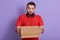 Delivery Concept. Portrait of curious caucasian handsome bearded delivery man wearing red casual t shirt, holding carton box