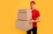 Delivery employee, male courier in red t-shirt and cap, with a bunch of cardboard boxes on yellow background