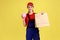 Delivery woman holding paper parcel and piggybank, looking at camera with happy facial expression.