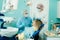 A dentist in a protective mask sits next to him and treats a patient in the dental office with an assistant