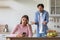 Depressed sad young caucasian lady with phone ignoring frustrated scolding man in kitchen interior