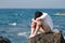 Depressed upset young Asian man covering face with hands sitting on the rock of sea shore