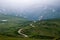 Descending winding mountain highway during fog and rain, around hills and green meadows.