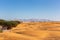 Desert landscape with sand dunes, residential buildings and wild Ghaf trees, with Hajar Mountains in the background, Al Madam