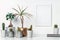 A desk by the white wall with lots of house plants, notebooks and a white mockup frame. A place for your text or graphics.
