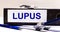 The desktop has a stethoscope, a blue pen, and a gray file folder with the text LUPUS. Medical concept