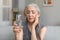 Despaired sad caucasian mature female with gray hair holds glass of water, suffers from pain