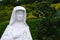 Detail of sculpture of polish Saint Faustyna Kowalska holding cross and rosary, decorative shrubs in background.