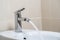 Details of white ceramic bidet with water running from tap in modern bathroom with beige tiles background. Close up