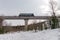 A diesel locomotive drives across a high concrete railway bridge over a snow-covered forest against a cloudy sky in winter