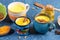 Different cups with golden turmeric milk and ingredients for its cooking on blue backdrop. Close-up, selective focus