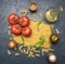 Different types of pasta, laid out with various ingredients, tomatoes, herbs, oil in a decanter seasonings on a dark rustic ba