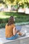 Digital addiction. Caucasian woman in white headphones sits on a parapet and hang out on the laptop in the public park