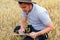 Digital farm. Portrait of farmer seating in gold wheat field and writing on tablet. Young man wearing cowboy hat in